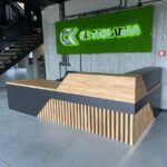 Moderní recepční pult s dřevěnou deskou a geometrickým čelem v industriálním interiéru
